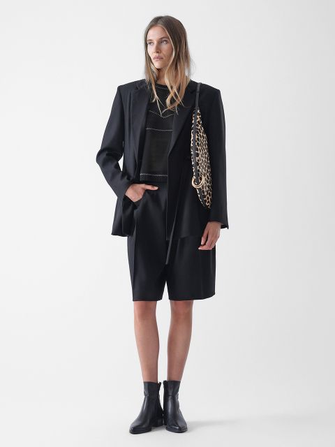 Eine Frau steht vor einem schlichten weißen Hintergrund und trägt einen schwarzen Blazer, schwarze knielange Shorts, ein schwarzes Oberteil, schwarze Ankle Boots und eine klobige Umhängetasche mit Goldkette. Sie hat langes, glattes Haar und einen neutralen Gesichtsausdruck.