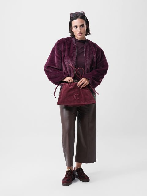 Eine Person steht vor einem schlichten weißen Hintergrund und trägt eine weinrote Kunstpelzjacke, eine braune Hose mit weitem Bein und weinrote Turnschuhe. Sie hält eine passende burgunderfarbene Handtasche und trägt eine Sonnenbrille auf dem Kopf.