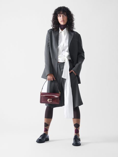 Eine Person mit lockigem Haar, die einen grauen übergroßen Blazer und dazu passende knielange Shorts, ein weißes Hemd mit langem Schal, eine dunkle Strumpfhose, gemusterte Socken und schwarze Halbschuhe trägt und eine weinrote Handtasche in der Hand hält, steht vor einem weißen Hintergrund.