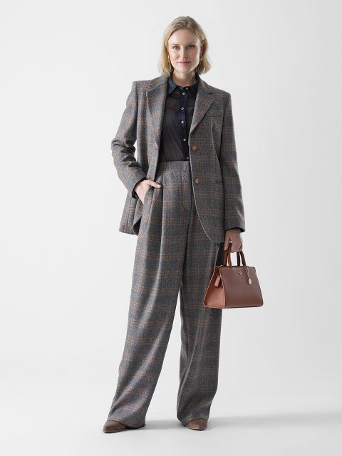 Eine Frau steht vor einem weißen Hintergrund und trägt einen grau karierten Anzug mit weit geschnittenen Hosen und einer schwarzen Bluse. Sie trägt eine braune Handtasche und hat hellblondes Haar, das einfach gestylt ist. Sie lächelt leicht.