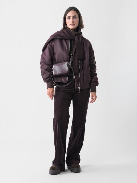 Eine Frau steht vor einem schlichten weißen Hintergrund und trägt eine dunkelbraune Jacke, eine passende Hose und einen über die Schultern drapierten Pullover. Sie trägt eine kleine Umhängetasche und hat dunkle Schuhe an.