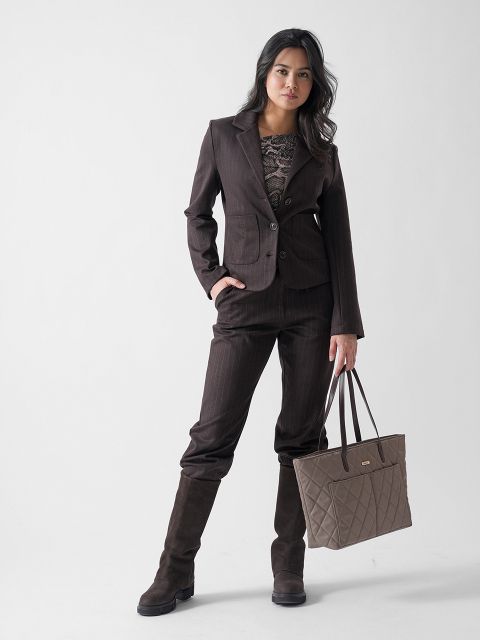 Eine Frau in einem dunkelbraunen Hosenanzug, einem gemusterten Oberteil und kniehohen Stiefeln steht vor einem schlichten Hintergrund und hält eine gesteppte taupefarbene Handtasche.