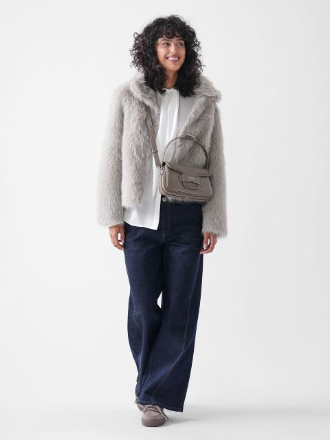 Eine Person mit lockigem Haar trägt eine hellgraue Kunstpelzjacke, eine weiße Bluse, dunkelblaue Jeans mit weitem Bein, taupefarbene Schuhe und eine passende taupefarbene Handtasche. Sie steht und lächelt vor einem schlichten weißen Hintergrund.