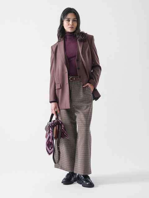 Eine Frau steht vor einem unifarbenen Hintergrund und trägt einen lilafarbenen Blazer mit Blumendetail, einen burgunderroten Rollkragenpullover, eine karierte Hose mit weitem Bein, schwarze Halbschuhe und einen passenden gemusterten Schal und eine Tasche.