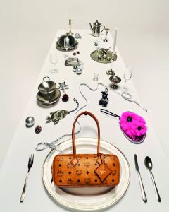 Ein langer weißer Tisch ist mit elegantem Silberbesteck, Teekannen und festlichen Dekorationen gedeckt. Eine hellbraune MCM-Handtasche steht auf einem Tablett im Vordergrund, und eine pinkfarbene Tasche liegt ebenfalls auf dem Tisch inmitten der Dekoration.
