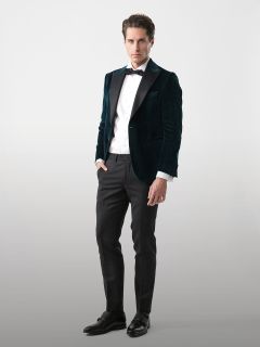 Mann mit dunkelgrüner Samtjacke, weißem Hemd, schwarzer Fliege, schwarzer Hose und schwarzen Schuhen vor einem hellen Hintergrund.