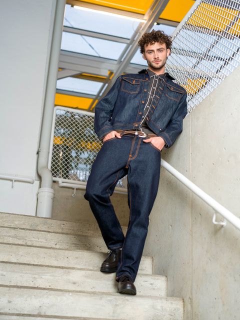 Ein Mann in dunkler Jeansjacke und Jeans steht auf einer Treppe, einen Fuß höher, und hält sich am Geländer fest, hinter ihm ein Maschendrahtzaun und ein gelbes Oberlicht.