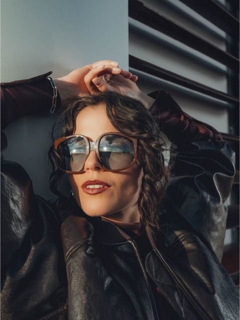 Eine Frau mit großer Sonnenbrille und schwarzer Lederjacke lehnt sich mit den Händen über dem Kopf zurück, das Sonnenlicht spiegelt sich in ihrer Brille.