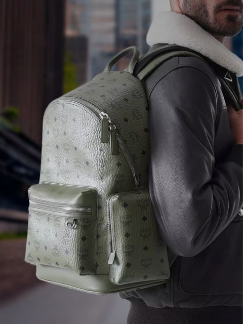 Eine Person in einer schwarzen Jacke trägt einen großen, strukturierten olivgrünen MCM-Rucksack mit Reißverschlusstaschen und steht im Freien in einer städtischen Umgebung.