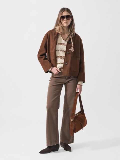 Frau, die eine braune Wildlederjacke, einen beige gemusterten Pullover, eine braune Hose, eine Sonnenbrille und braune Schuhe trägt und eine braune Wildledertasche mit sich führt, steht vor einem weißen Hintergrund.