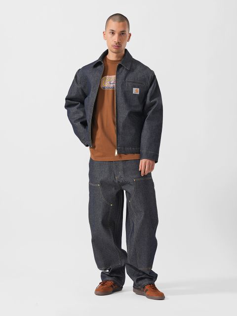 Ein Mann, der eine lockere dunkle Jeansjacke und Jeans, ein braunes grafisches T-Shirt und braune Schuhe trägt, steht vor einem schlichten weißen Hintergrund.