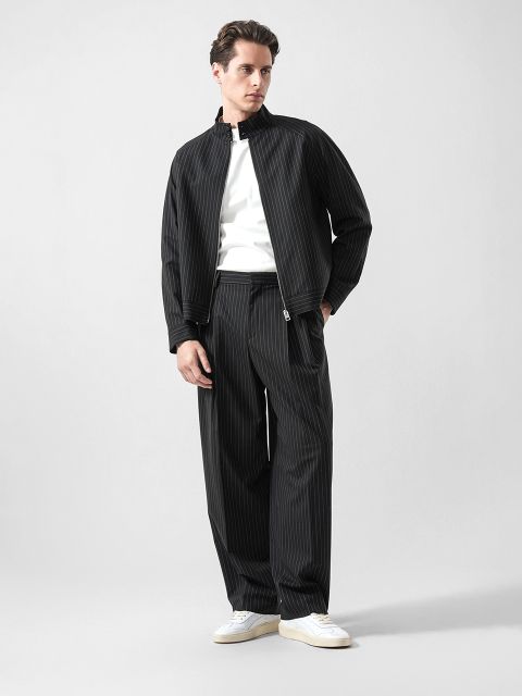 Ein Mann, der eine schwarze Nadelstreifenjacke und -hose über einem weißen Hemd mit weißen Turnschuhen trägt, steht vor einem schlichten hellen Hintergrund.