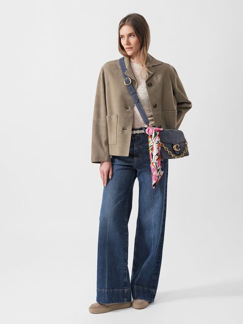 Frau in taupefarbener Jacke, spitzenbesetztem Oberteil, Jeans mit weitem Bein und Halbschuhen, die eine Umhängetasche mit einem bunten Schal in der Hand hält, steht vor einem weißen Hintergrund.