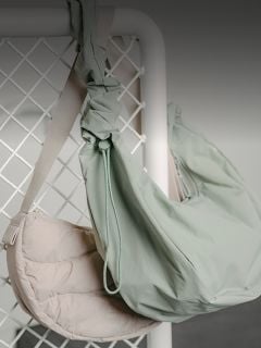 Zwei Umhängetaschen, eine hellbeige und eine blassgrüne, hängen an einem weißen Metallgitter vor einem neutralen Hintergrund.