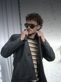 Mann mit lockigem Haar, der eine Sonnenbrille, einen gestreiften Pullover und eine schwarze Lederjacke trägt und seinen Kragen vor einer weißen Ziegelwand zurechtrückt.