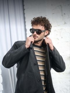 Ein Mann mit lockigem Haar, der eine Sonnenbrille, eine schwarze Lederjacke und einen gemusterten Pullover trägt, steht vor einer weißen Backsteinmauer und rückt seinen Kragen zurecht.