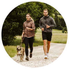 Eine Frau und ein Mann joggen an einem bewölkten Tag im Freien auf einem Kiesweg mit einem Hund, umgeben von Grün und Bäumen.