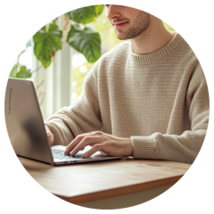 Mann im beigen Pullover, der an einem Holztisch auf einem Laptop tippt, mit Grünpflanzen und natürlichem Licht im Hintergrund.