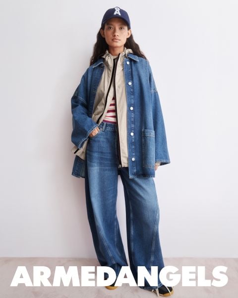 Eine Person in Jeans mit weitem Bein, Jeansjacke, gestreiftem Hemd, Mütze und Turnschuhen steht vor einer weißen Wand. Am unteren Rand steht "ARMEDANGELS" geschrieben.