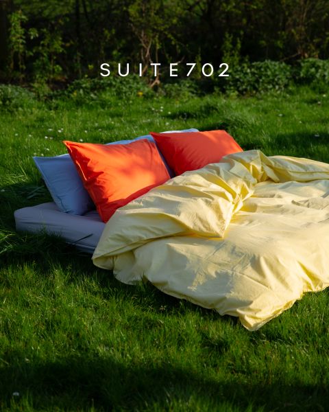 Ein Bett mit roten und weißen Kissen und gelbem Bettzeug steht im Freien auf grünem Gras im Sonnenlicht. Darüber erscheint der Text "SUITE702".