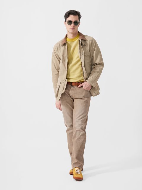 Ein Mann mit Sonnenbrille, beiger Jacke, gelbem Pullover, hellbrauner Hose und gelben Turnschuhen steht selbstbewusst vor einem schlichten weißen Hintergrund.