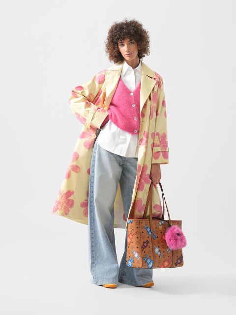 Frau in gelbem Trenchcoat mit Blumenmuster, rosa Weste, weißem Hemd und Jeans mit weitem Bein, die eine braune Tragetasche mit rosa Pompon in der Hand hält, steht vor einem weißen Hintergrund.