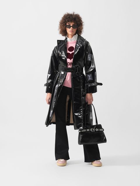 Eine Frau mit Sonnenbrille trägt einen glänzenden schwarzen Trenchcoat, ein rosa-weißes Hemd, eine schwarze Hose und hält eine schwarze Handtasche. Sie steht vor einem schlichten Hintergrund.