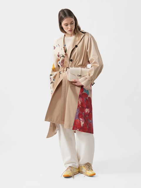 Frau in beigem Trenchcoat mit Blumenmuster, weißer Hose, gelben Turnschuhen und einer cremefarbenen Tasche vor weißem Hintergrund.