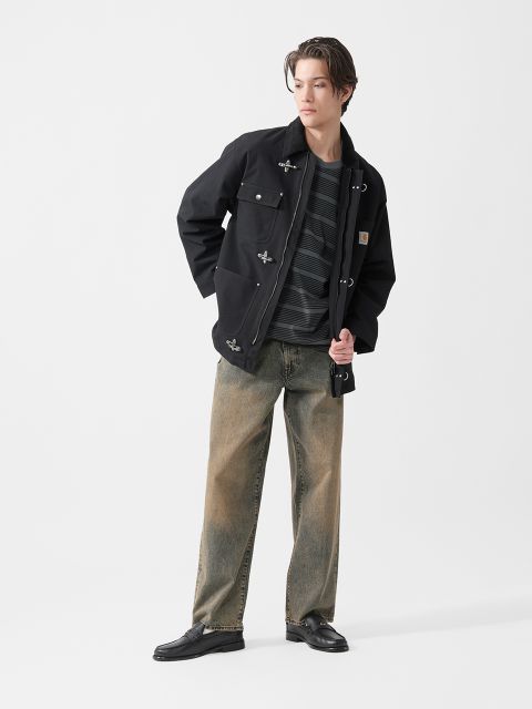 Ein junger Mann in schwarzer Jacke, gestreiftem Hemd, weiten, verwaschenen Jeans und schwarzen Schuhen steht vor einem schlichten weißen Hintergrund und schaut zur Seite.