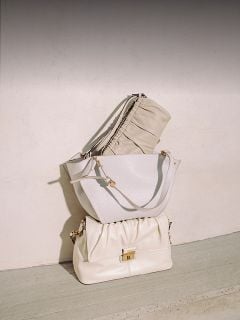 Drei elegante beigefarbene und weiße Handtaschen sind vor einer hellen Wand und einem Betonboden gestapelt und im natürlichen Sonnenlicht ordentlich angeordnet.