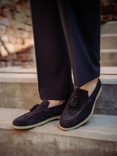 Person in dunklen Wildlederschuhen mit Quasten und dunkler Hose, stehend auf einer Betontreppe im Freien mit einem unscharfen Backsteinmauer-Hintergrund.