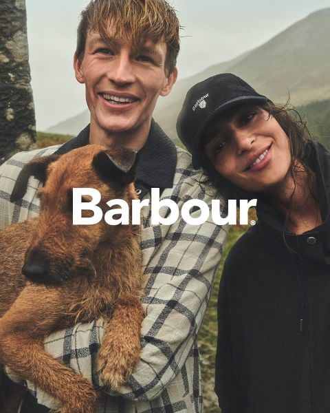 Ein lächelnder Mann, der einen braunen Hund hält, steht neben einer lächelnden Frau mit einer schwarzen Mütze im Freien. Das Wort "Barbour" ist auf dem Bild eingeblendet.