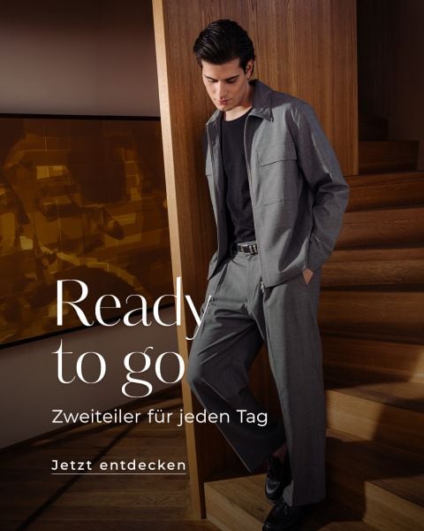 Ein Mann in einem grauen Anzug und schwarzem Hemd steht auf einer Holztreppe. Der Text lautet: "Bereit zu gehen. Zweiteiler für jeden Tag. Jetzt entdecken.