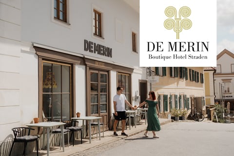 Ein Paar hält sich vor dem De Merin Boutique Hotel Straden mit Sitzgelegenheiten im Freien und sonnigen Gebäuden an den Händen. Das Hotellogo erscheint oben rechts.