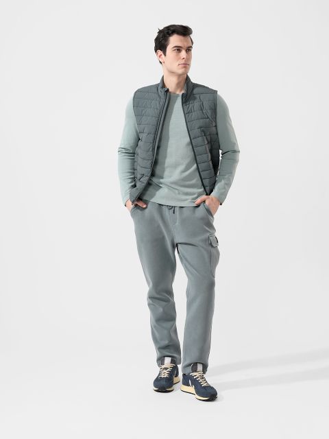 Ein Mann, der eine graue Steppweste, ein hellgrünes langärmeliges Hemd, graue Jogginghosen und blaue Turnschuhe trägt, steht vor einem schlichten weißen Hintergrund.