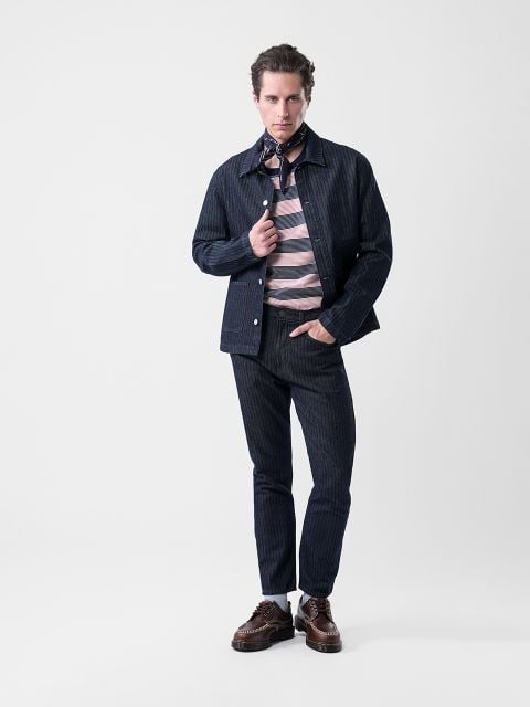 Ein Mann posiert in einer marineblau gestreiften Jacke und Hose, einem rosa und grau gestreiften Hemd, braunen Schuhen und einem gemusterten Schal vor einem schlichten weißen Hintergrund.