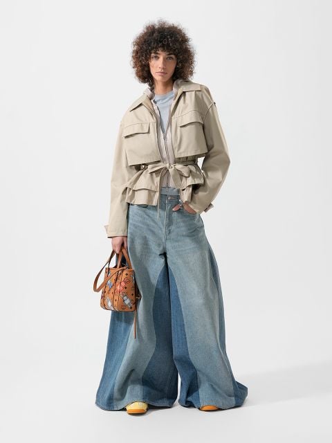 Eine Person, die eine beigefarbene Jacke, weit geschnittene blaue Jeans und gelbe Schuhe trägt und eine braun gemusterte Handtasche in der Hand hält, steht vor einem weißen Hintergrund.