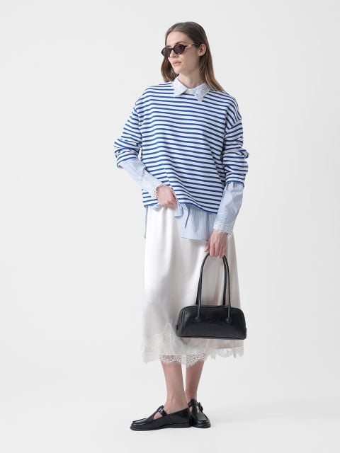 Frau mit Sonnenbrille, die einen blau-weiß gestreiften Pullover über einem Hemd mit Kragen, einen weißen Rock mit Spitzenbesatz, schwarze Halbschuhe und eine schwarze Handtasche trägt.