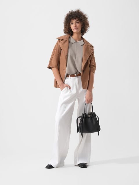 Frau mit lockigem Haar, die eine hellbraune Jacke, ein beiges Hemd und eine weiße Hose mit weitem Bein trägt und eine schwarze Handtasche in der Hand hält, steht vor einem schlichten Hintergrund.