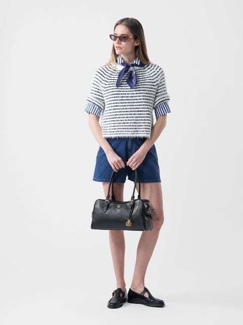 Frau mit Sonnenbrille, die einen gestreiften Pullover über einem Hemd mit Kragen, Jeansshorts und schwarze Schuhe trägt und eine schwarze Handtasche vor einem weißen Hintergrund hält.