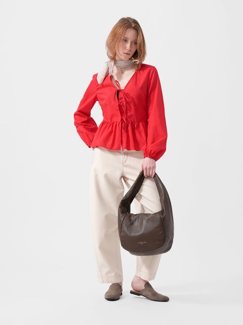 Frau mit roter Bluse, cremefarbener Hose mit weitem Bein, braunen Flats, Schal und einer großen braunen Handtasche in der Hand, vor einem schlichten weißen Hintergrund.