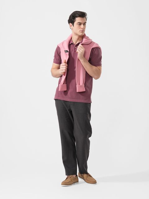 Mann in Freizeitkleidung, mit kastanienbraunem Polohemd, dunkler Hose, braunen Schuhen und einem über die Schultern drapierten rosa Pullover vor weißem Hintergrund.