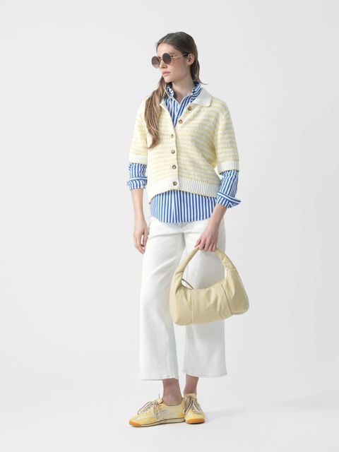 Frau mit Sonnenbrille, gestreiftem blauen Hemd, gestreifter Strickjacke, weißer Hose und gelben Schuhen hält eine hellgelbe Handtasche und steht vor einem weißen Hintergrund.