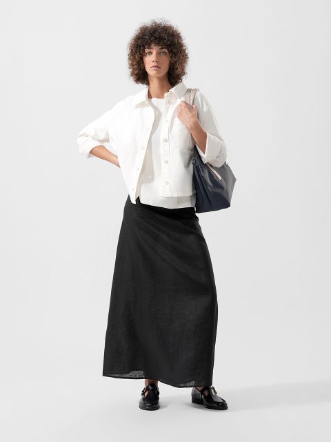 Eine Frau steht selbstbewusst da, trägt eine weiße Jacke, einen schwarzen Maxirock, schwarze Halbschuhe und eine große schwarze Tragetasche vor einem schlichten Hintergrund.