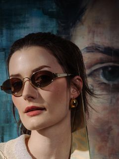 Frau mit braunem Haar, die eine ovale Sonnenbrille und einen goldenen Ohrring trägt, steht vor einem gemalten Porträt mit einem großen Auge und blauen Farbtönen.