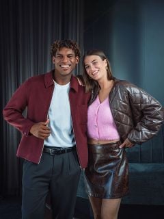 Ein lächelnder Mann in einer burgunderroten Jacke steht neben einer Frau in einem rosa Oberteil und einem braunen Rock; beide posieren in einem Innenraum vor einem dunklen Hintergrund.