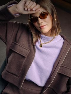Eine Frau mit brauner Sonnenbrille, Jacke und lavendelfarbenem Pullover stützt ihren Arm auf die Stirn; sie trägt Goldschmuck und hat einen selbstbewussten Gesichtsausdruck.