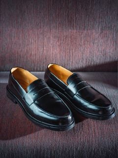 Ein Paar glänzende schwarze Leder-Loafer, die nebeneinander auf einer strukturierten, weinroten Stoffunterlage liegen.