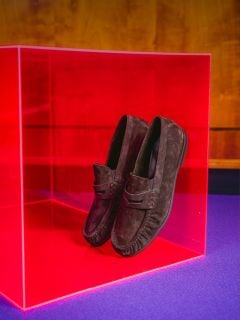 Braune Wildleder-Loafer, präsentiert in einem durchsichtigen rosa Würfel, vor einem Holzhintergrund und auf einer violetten Unterlage.