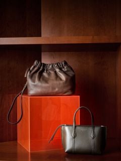 Zwei elegante Lederhandtaschen, eine braune und eine olivgrüne, präsentiert auf einem roten Würfelregal vor einem Holzhintergrund.